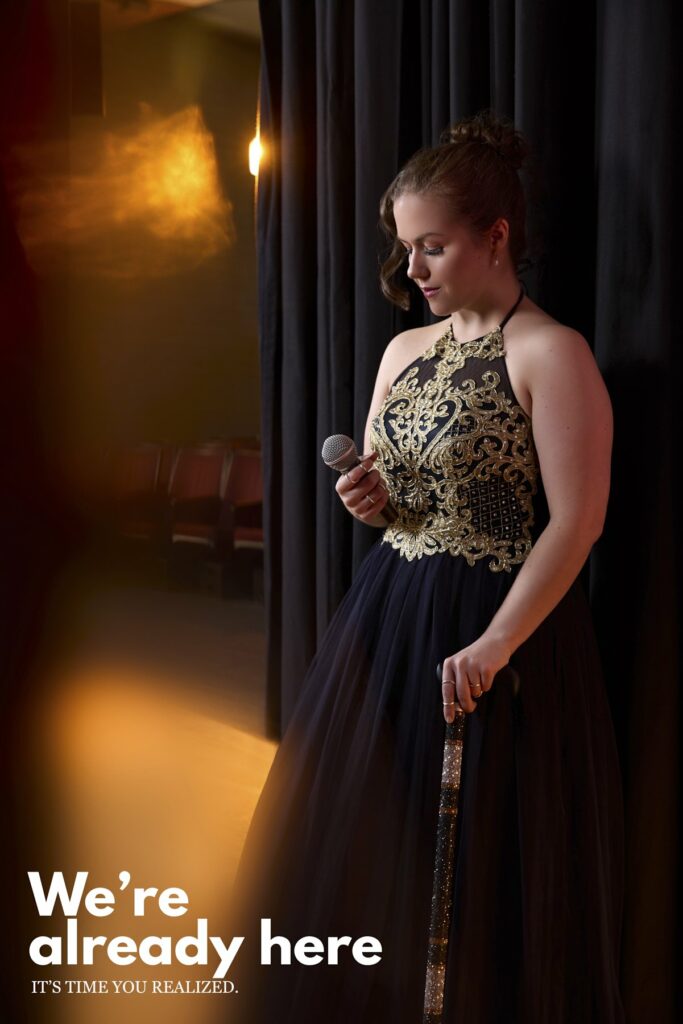 An elegant performer with brown hair in a golden embroidered gown takes a moment backstage. A microphone is in her right hand and she holds a cane with her left hand. A text says: we're already here, it's time you realized.
