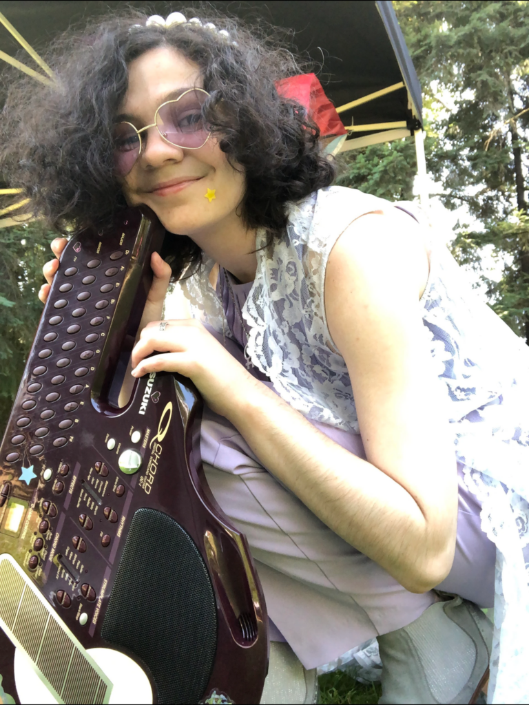 A white non-binary person with black curly short hair and pink-tinted glasses holding a musical instrument that resembles an autoharp.