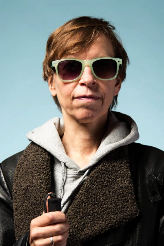A white woman with short brown hair wearing sunglasses with a green frame and a brown and grey hoodie. She holds a white cane in her right hand.