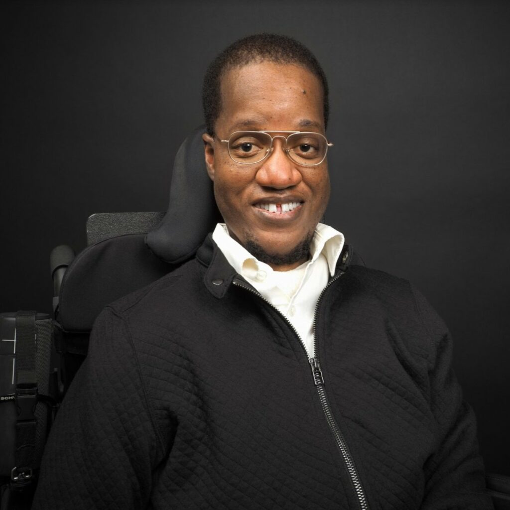 A black man with grey metallic framed glasses wearing a white shirt and a black jacket.