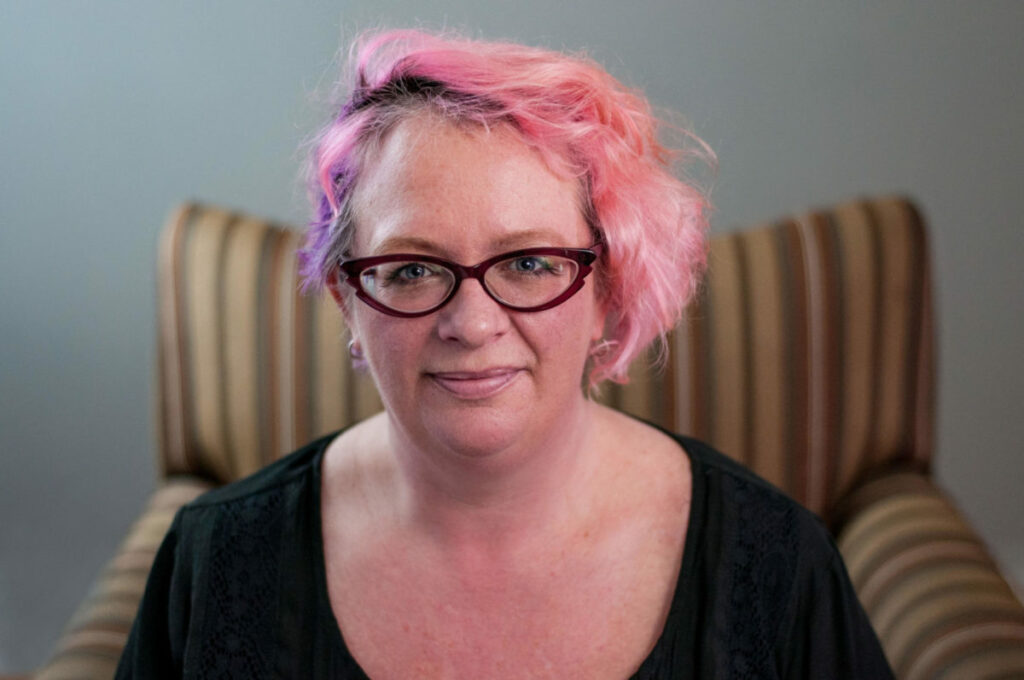 A white woman with pink short hair and dark red glasses wearing a black top. She is sitting in an armchair with yellow, green and brown stripes.