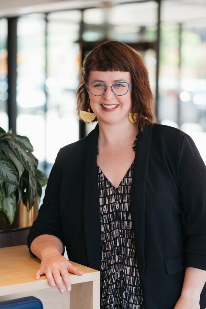 A white woman with dark red hair wearing glasses with blue frames and semi circular golden earring. She wars a black cardigan over a black and white top.