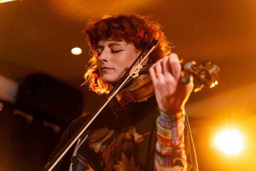 A white woman with red hair playing the violin on a stage lit by a bright orange light; her eyes are closed while she is playing.