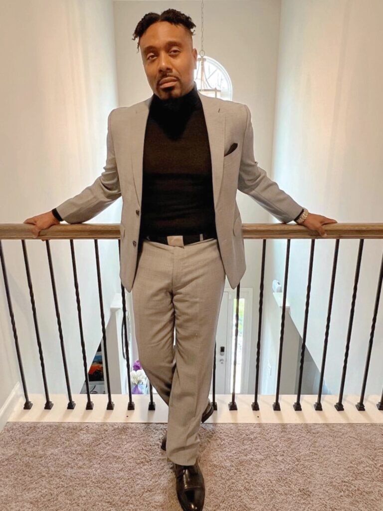 A black man wearing a beige suit jacket over a black top and beige suit pants stands with his hands on a staircase handrail.