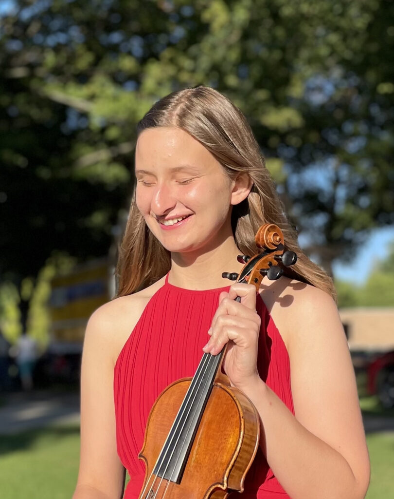 A white woman with long brown hair wearing a red dress, holding her violin; her eyes are closed.