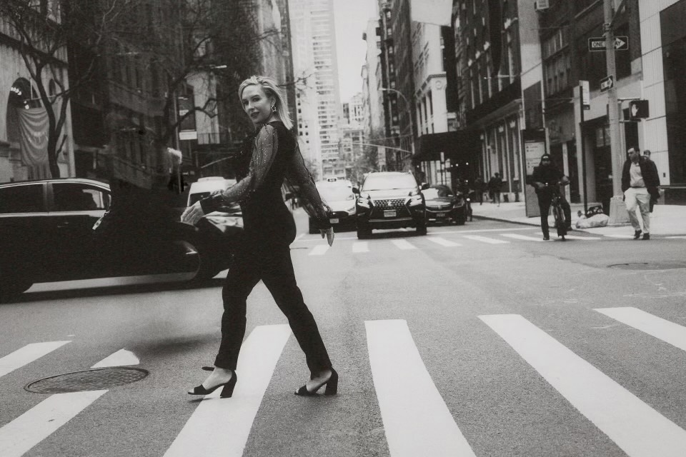 A white woman with long hair, shiny top and black pants, crossing the street on a crosswalk. Behind her are cars. It is a black and white picture taken in the streets of New York City.