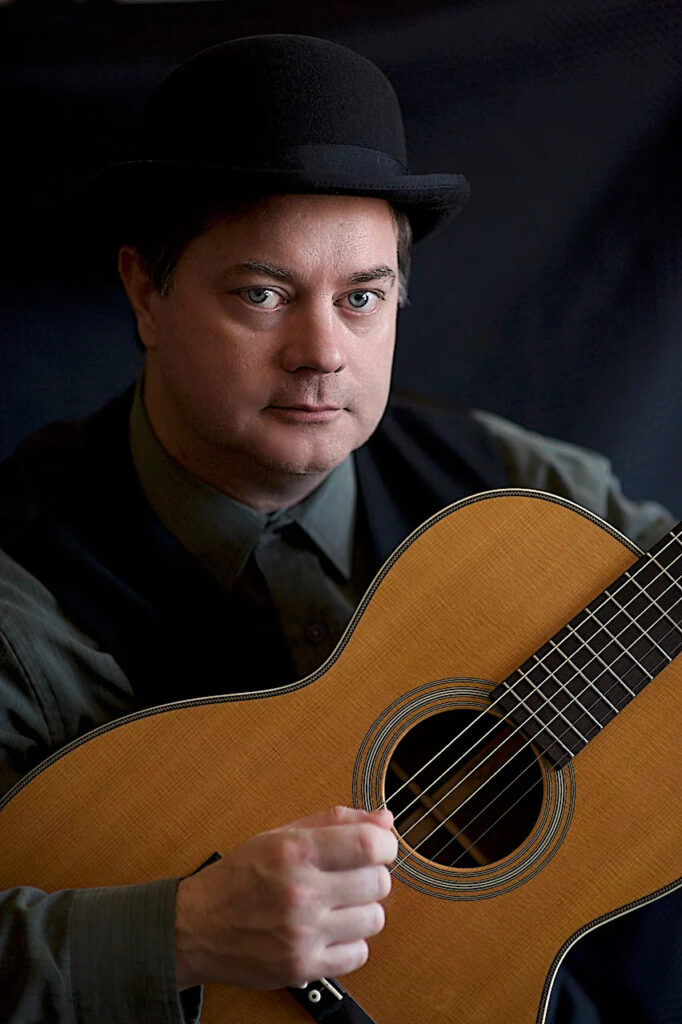 Un homme blanc aux yeux bleus portant un chapeau noir, une chemise grise et un gilet sans manche noir, tenant une guitare.