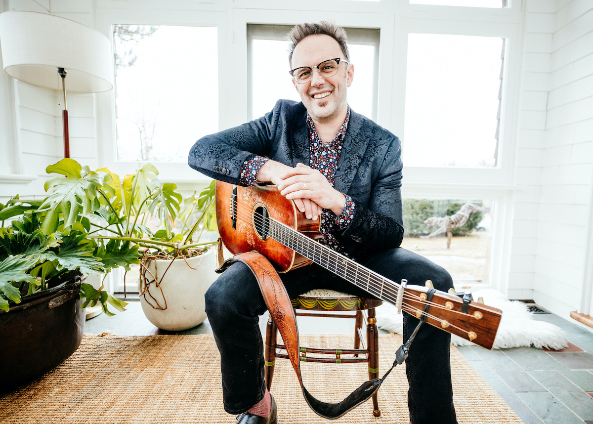 A white man with glasses wearing a grey jacket over a black shirt with flowers motifs. He sits on a chair and holds a guitar. Behind him are plants.