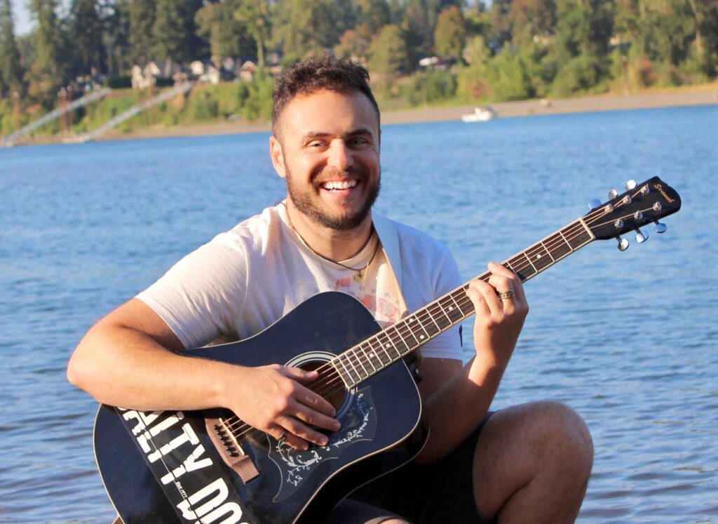 Un homme blanc aux cheveux courts portant un short noir et un t-shirt blanc. Il tient une guitare. Il est assis devant un lac.