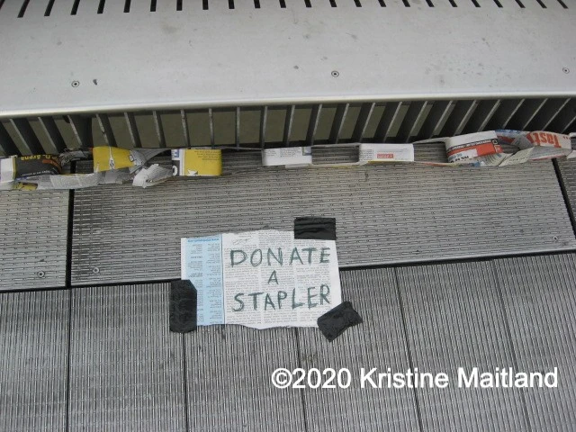 A paper sign posted on the ground that says "Donate a stapler". There is a paper chain above the sign.