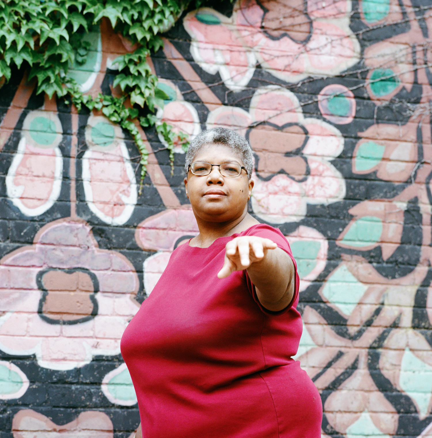 A black woman with a short afro and glasses wearing a mauve t-shirt dress standing in front of a tree brick wall. Her hand is in front of her, toward the camera.