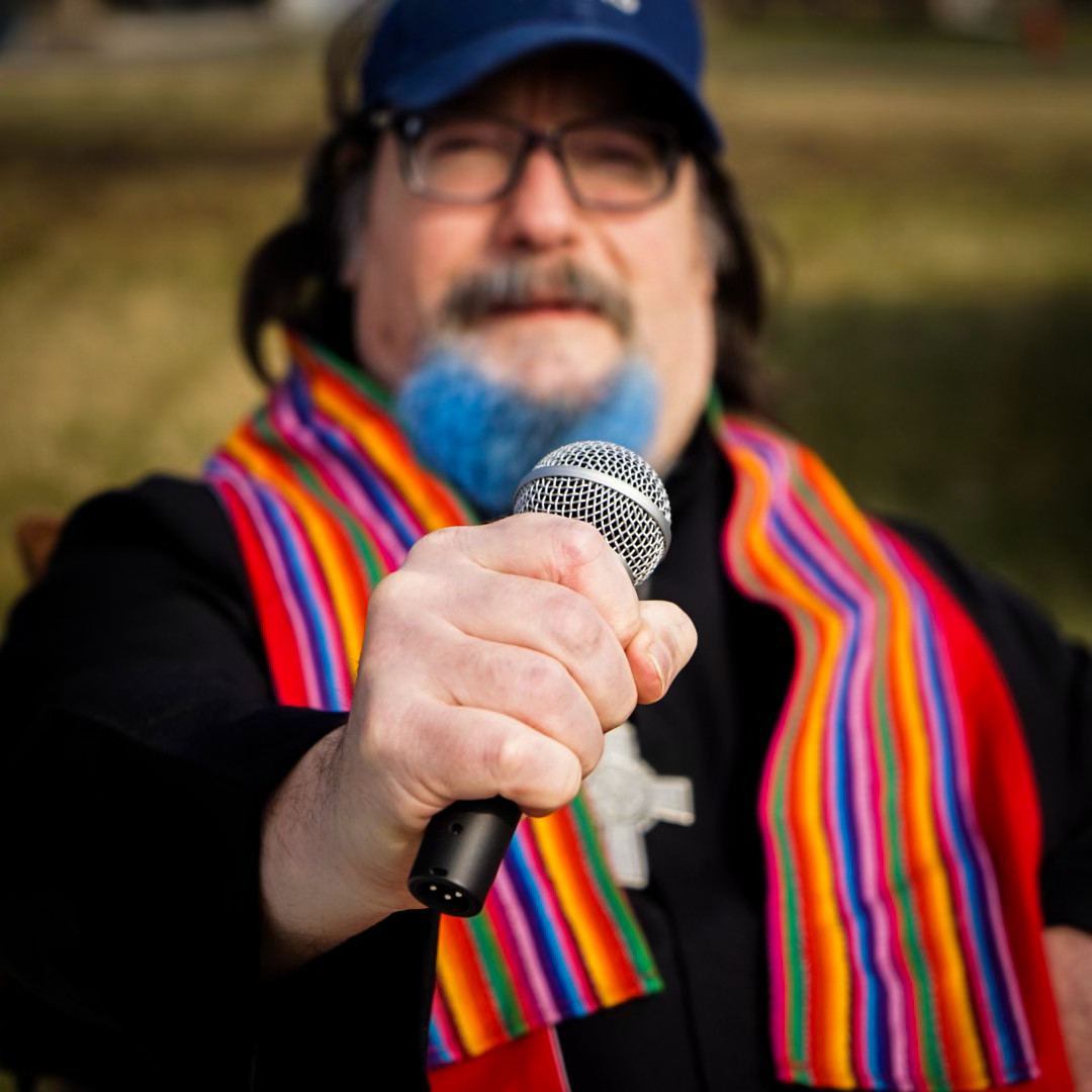 Un homme blanc avec des lunettes, une barbe bleue, une casquette bleue et une écharpe multicolore tient un micro.