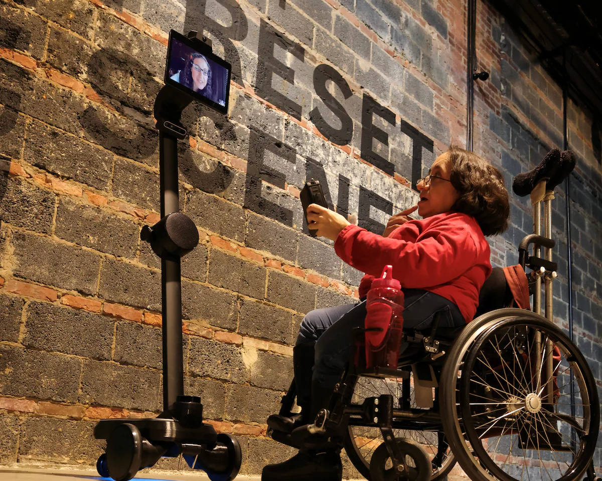 The face of a white woman with dark hair wearing glasses is displayed on the computer screen of a robot called Lisbot. She dialogues with another white woman with brown hair wearing glasses who sits in a wheelchair, behind her wheelchair are crutches, and a red water bottle is attached to the frame of her wheelchair.