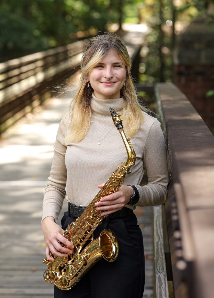 Une femme blanche aux longs cheveux blonds, vêtue d'un haut beige et d'un pantalon noir, debout, tenant son saxophone alto.