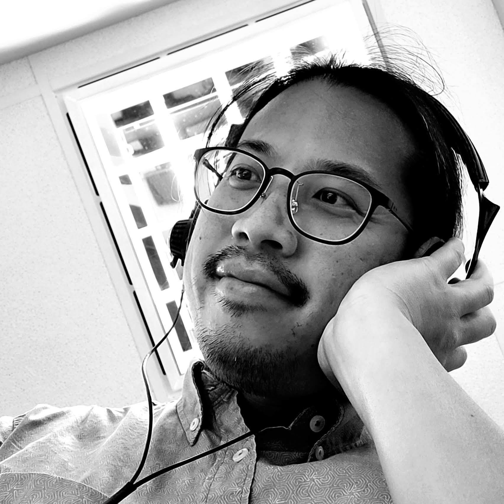 A black and white picture of a South-East Asian man with glasses and headphones. He has a light moustache and a light beard, and wears a shirt with geometrical patterns.