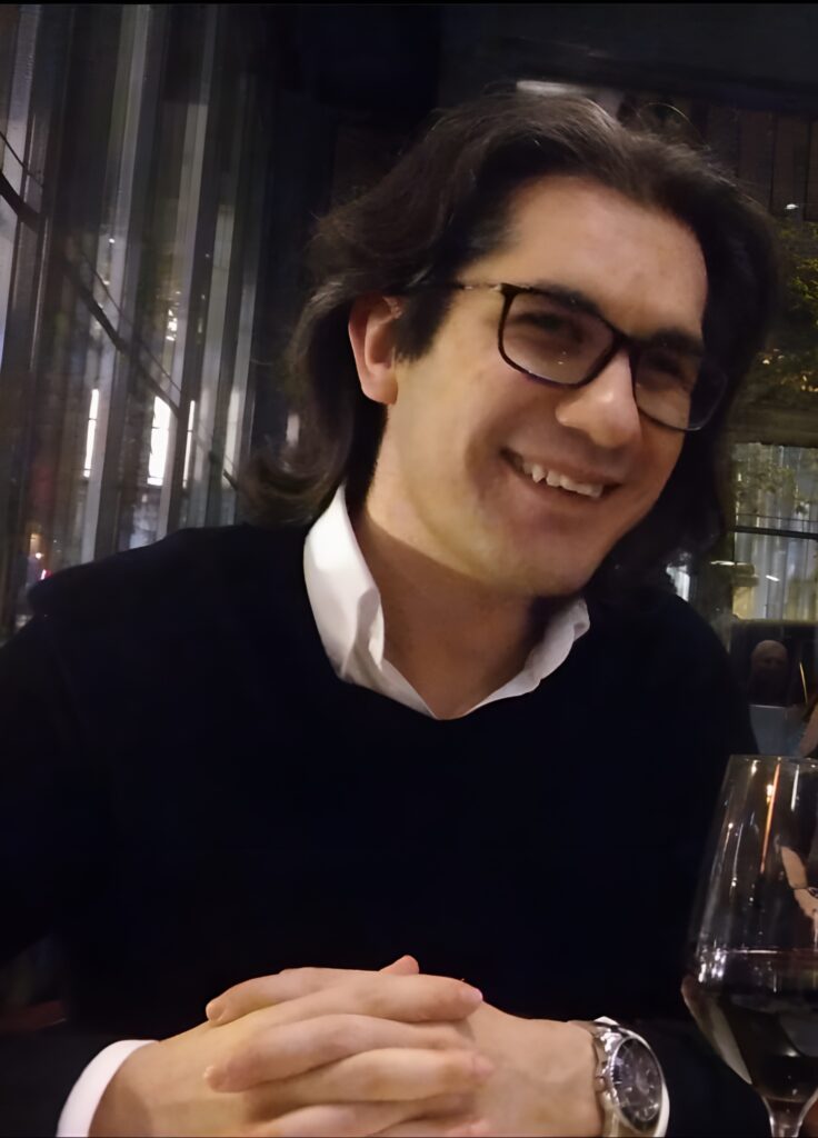 A white man with dark shoulder length hair and dark-rimmed glasses, wearing a black sweater over a white shirt, smiling at the camera.