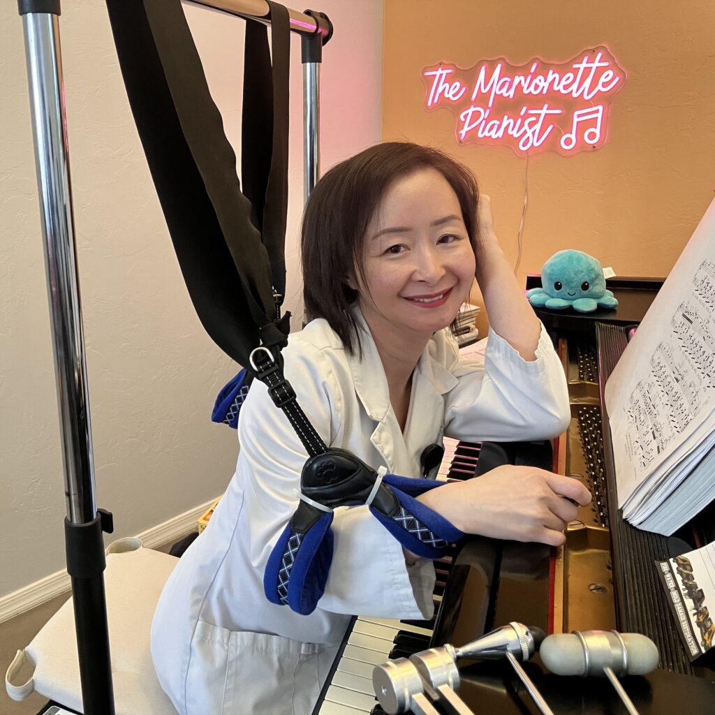 An East-Asian woman wearing a white jacket and white pants, sitting at the piano with a score open in front of her. A metallic frame is installed around her, straps attached to the frame are holding her arms. The inscription "The Marionette Pianist" is written in pink on the wall.