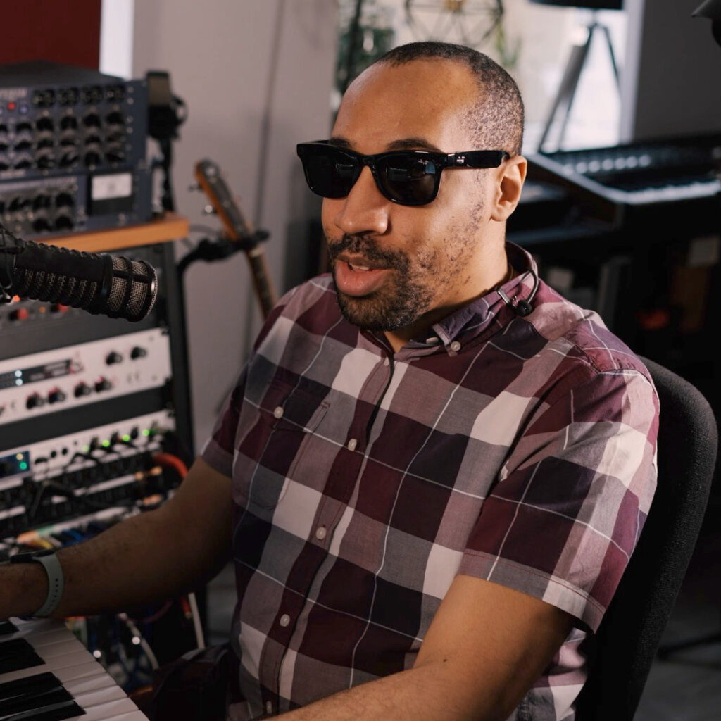 A black man with short hair and short beard, wearing sunglasses and a check shirt. In front of him, a microphone and a piano keyboard. Other audio equipment is surrounding him.