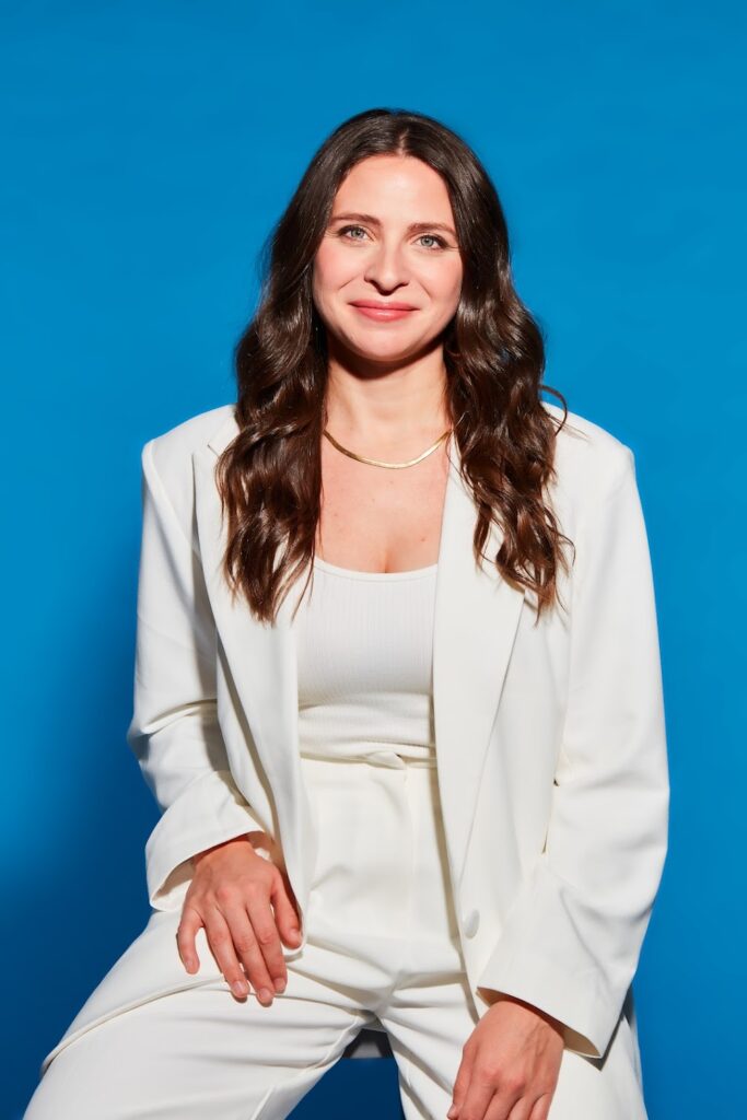 A white woman with brown curly hair wearing a golden necklace, dressed in a white jacket over a white top and what pants. The background is entirely blue.