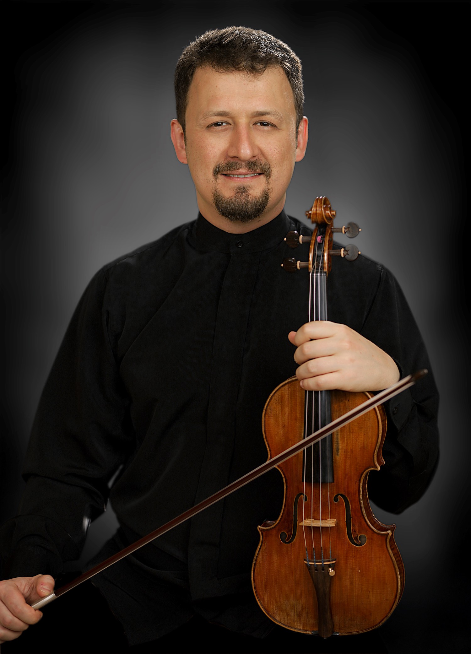 A white man with a short beard and moustache wearing a black top and holding a violin.