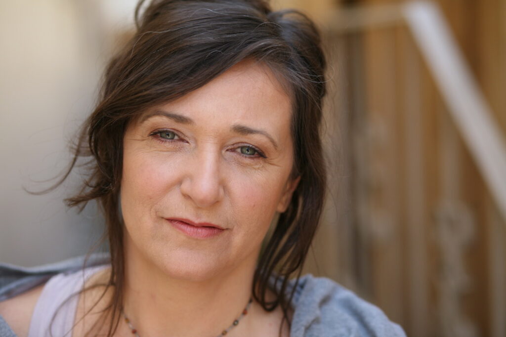 A white woman with brown hair and blue eyes wearing a blue cardigan over a purple top.